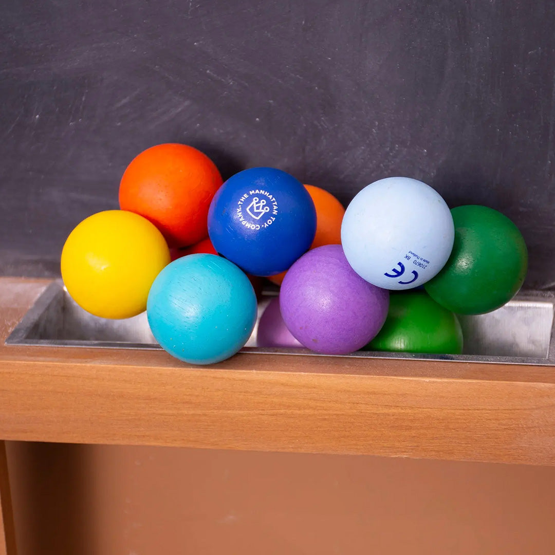 Manhattan Toy babyspeelgoed, een veelkleurige houten rammelaar van verbonden kralen liggend op een houten plank voor een schoolbord.