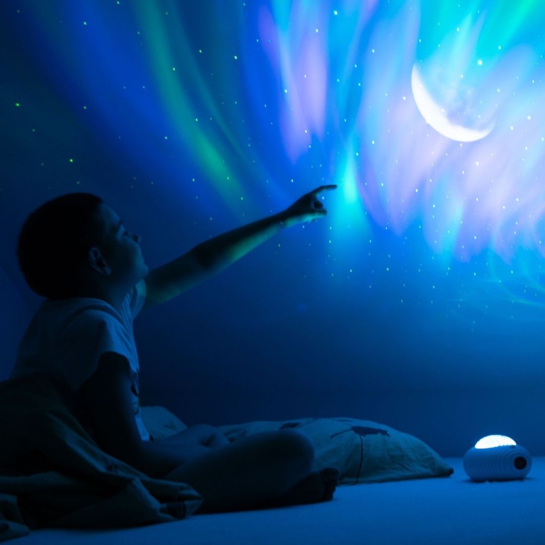 Kind wijst naar een muur verlicht met blauwe en groene aurora, sterren en een maan, geprojecteerd door de mary's lichtprojector.
