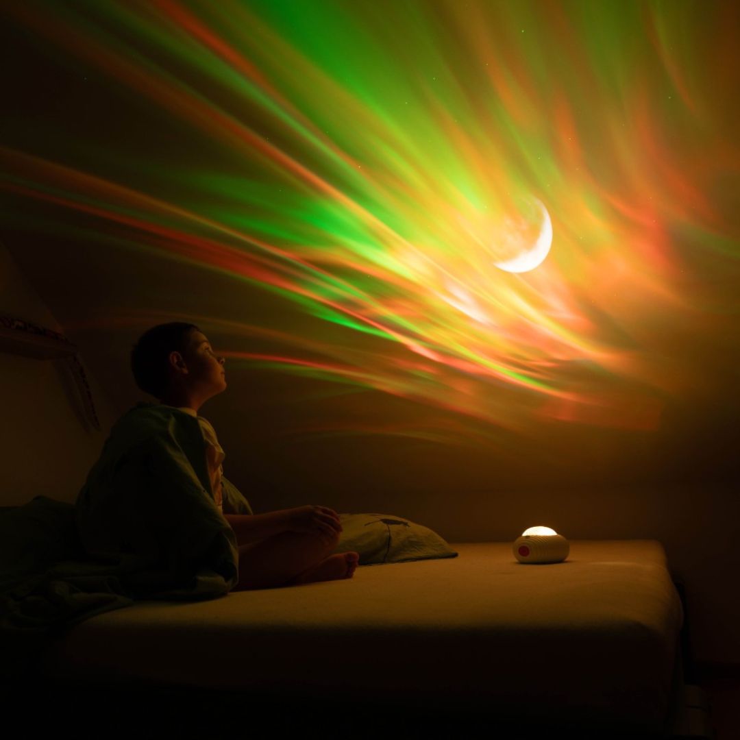 Kind kijkt naar een muur verlicht met rode en groene aurora, sterren en een maan, geprojecteerd door de mary's lichtprojector.