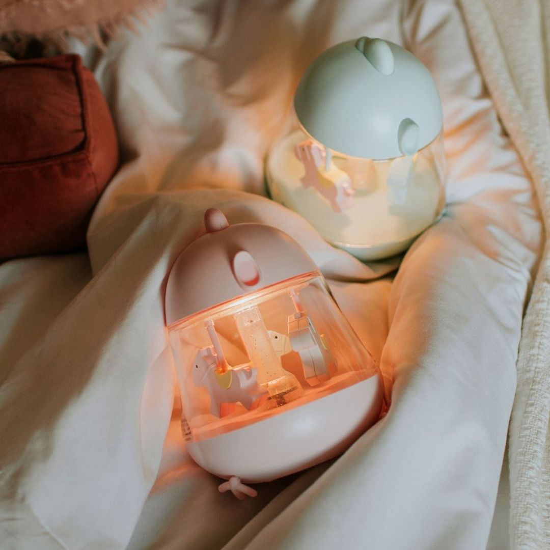 Twee Mary's muziekdozen met nachtlampje, een roze en een mintgroene, liggen verlicht op een witte deken.