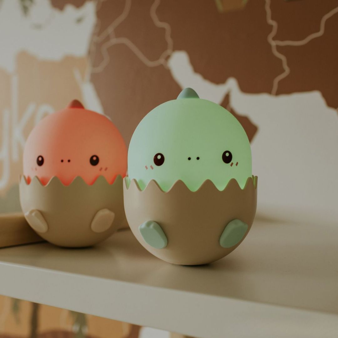Twee oplichtende baby dino nachtlampjes in eierschalen, één oranje en één groen, op een plank.
