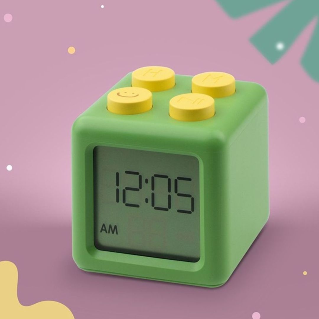 Mary's groene kubusvormige digitale wekker met gele knoppen toont 12:05 AM tegen een roze achtergrond.