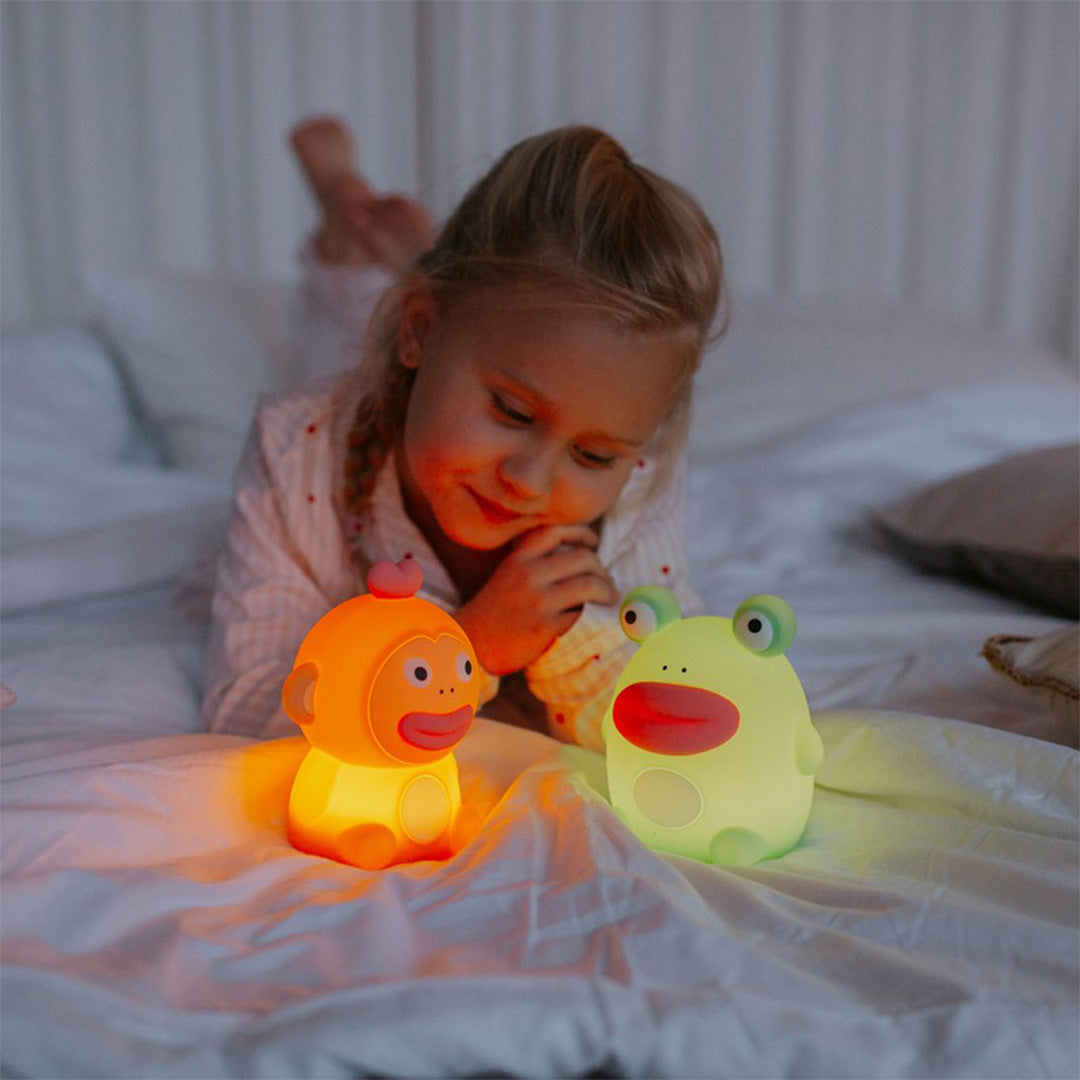 Een lachend meisje in bed kijkt naar een oplichtend Mary's kikker nachtlampje en een aap nachtlampje.