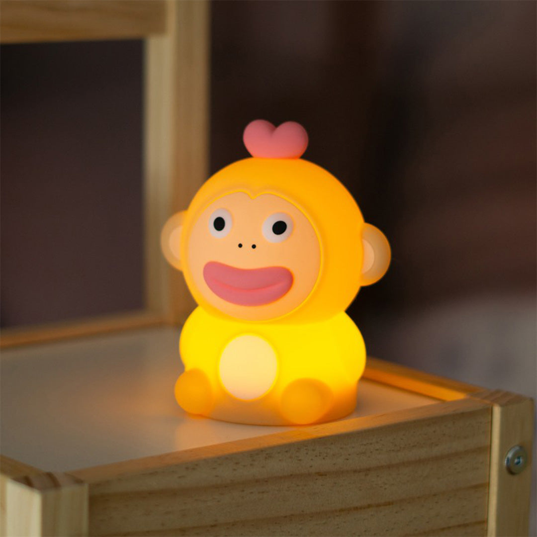 Mary's nachtlampje in de vorm van een aapje dat warm geel licht geeft, staand op een houten plank.