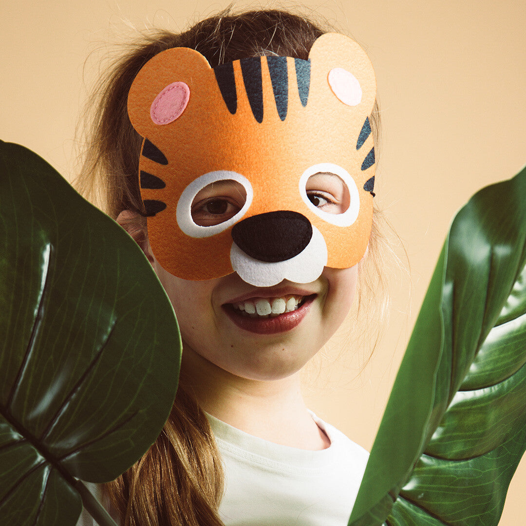 Een lachend kind draagt het tijgermasker tussen groene bladeren.