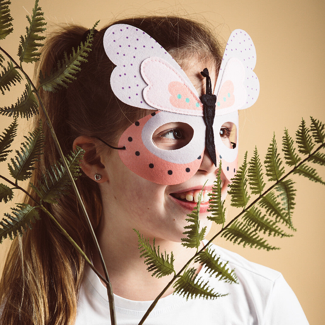 Een kind draagt het Folat vilten vlindermasker en kijkt opzij, met groene varens op de voorgrond.