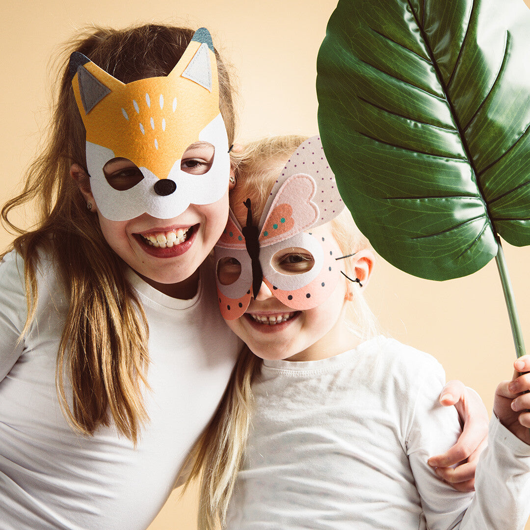 Twee lachende kinderen dragen een Folat vilt vossenmasker en een vlindermasker, met een groot groen blad.