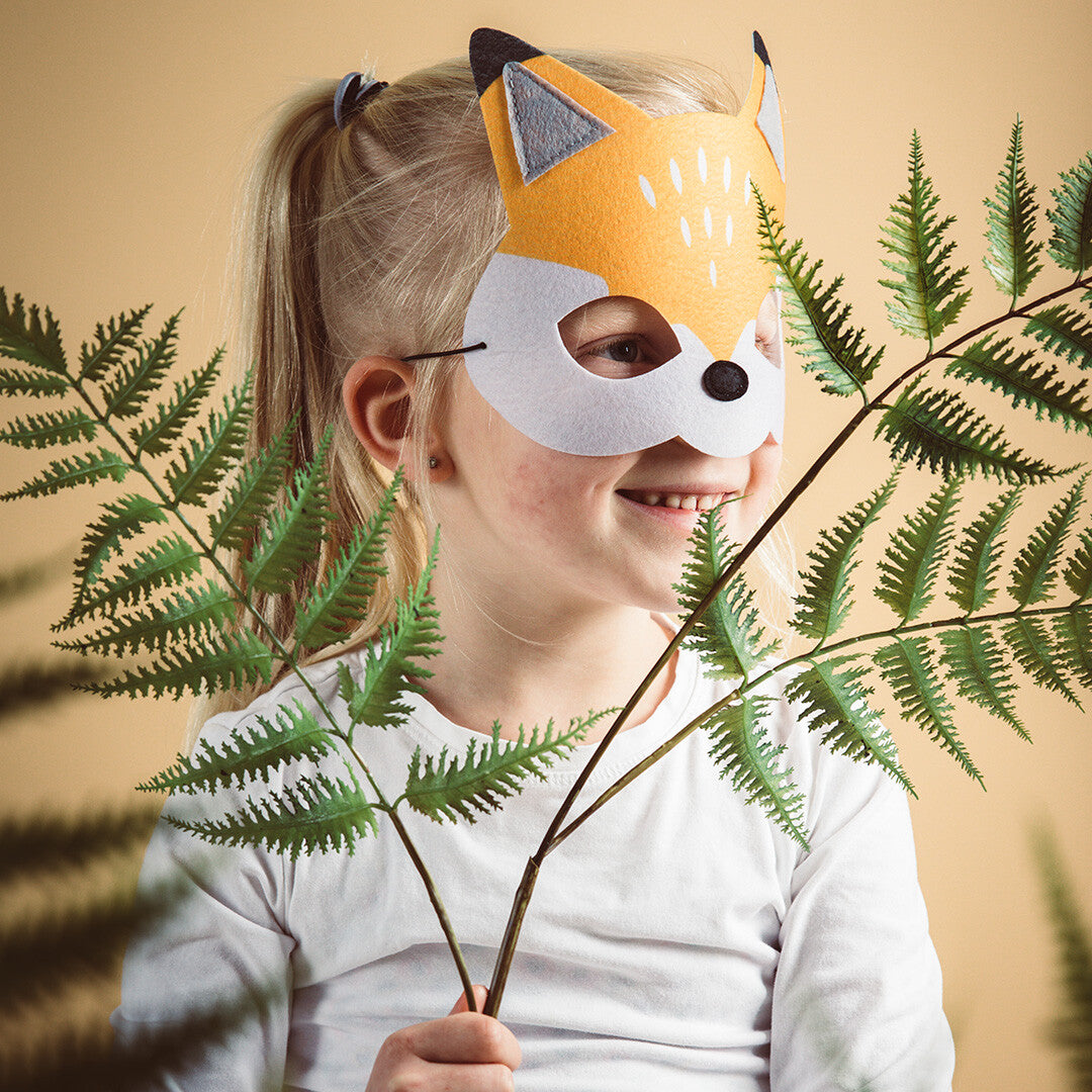 Een kind draagt een Folat vilt vossenmasker en houdt groene varenbladeren vast.