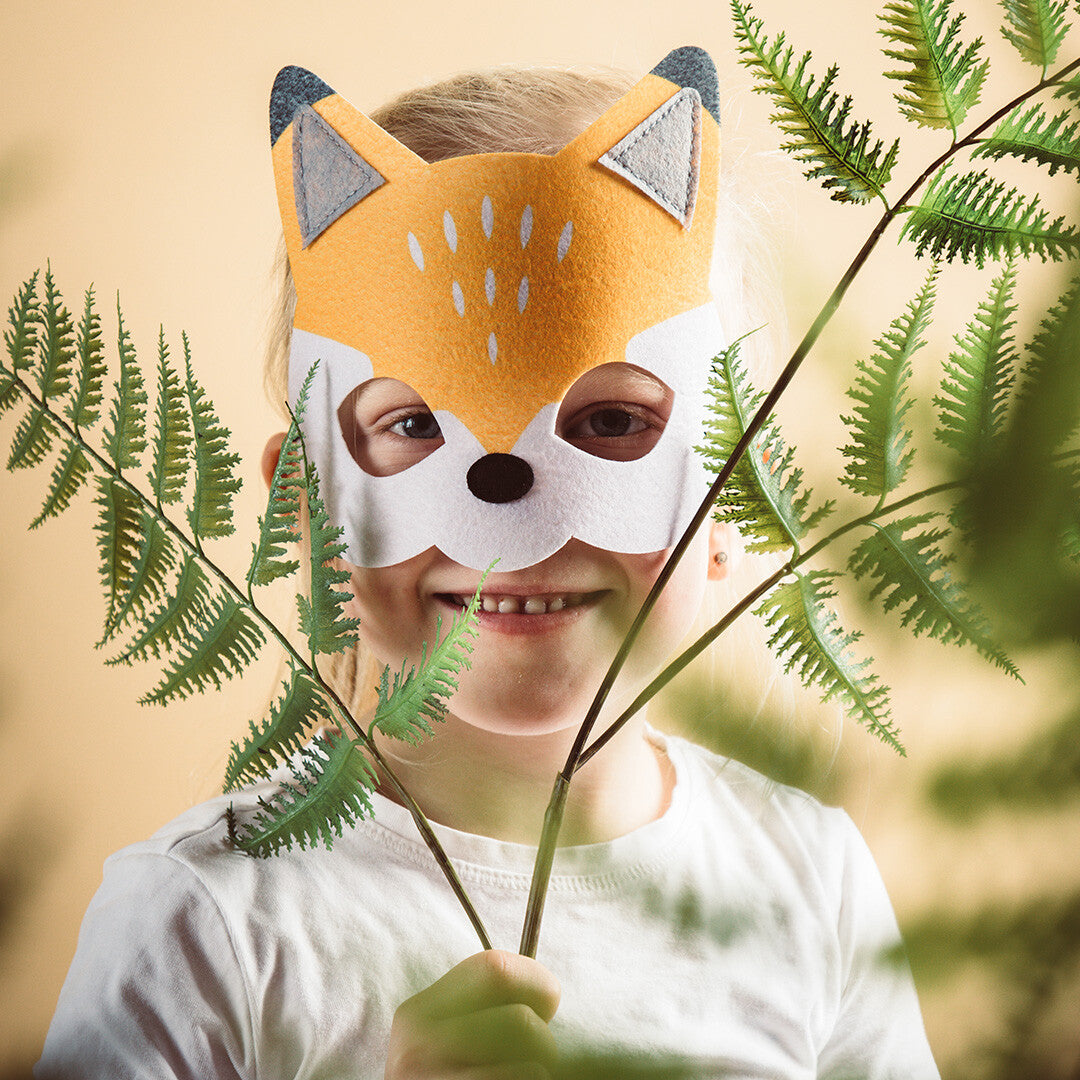 Een kind draagt een Folat vilt vossenmasker en lacht achter groene varenbladeren.
