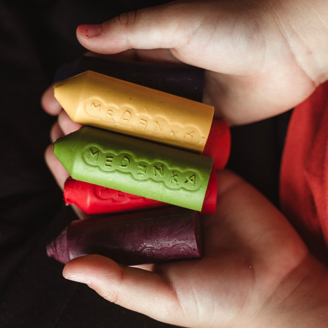 Kinderhanden houden een stapel Medenka bijenwaskrijtjes in verschillende kleuren vast.