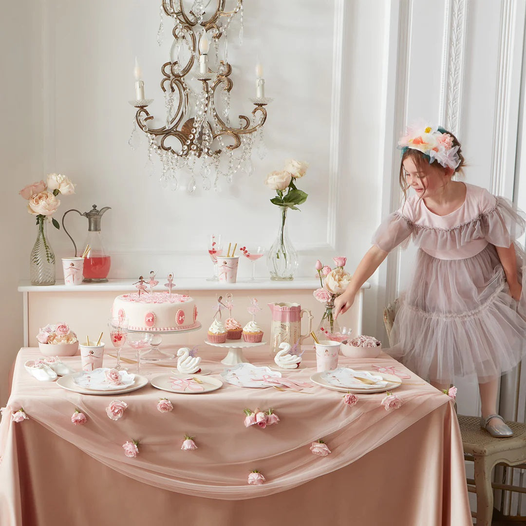 Een meisje in een roze jurk bij een feestelijk gedekte tafel met roze decoraties, een taart en feestartikelen.