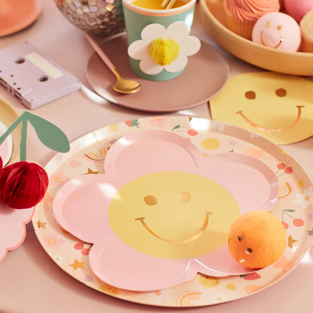 Meri Meri roze papieren feestbordjes met gele smiley gezichten op een dienblad tussen andere feestartikelen.