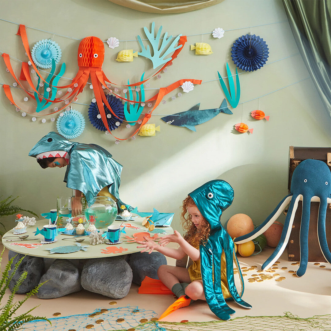 Twee kinderen in haai- en octopuskostuum aan een feestelijk gedekte tafel met Meri Meri onderwater thema decoraties.