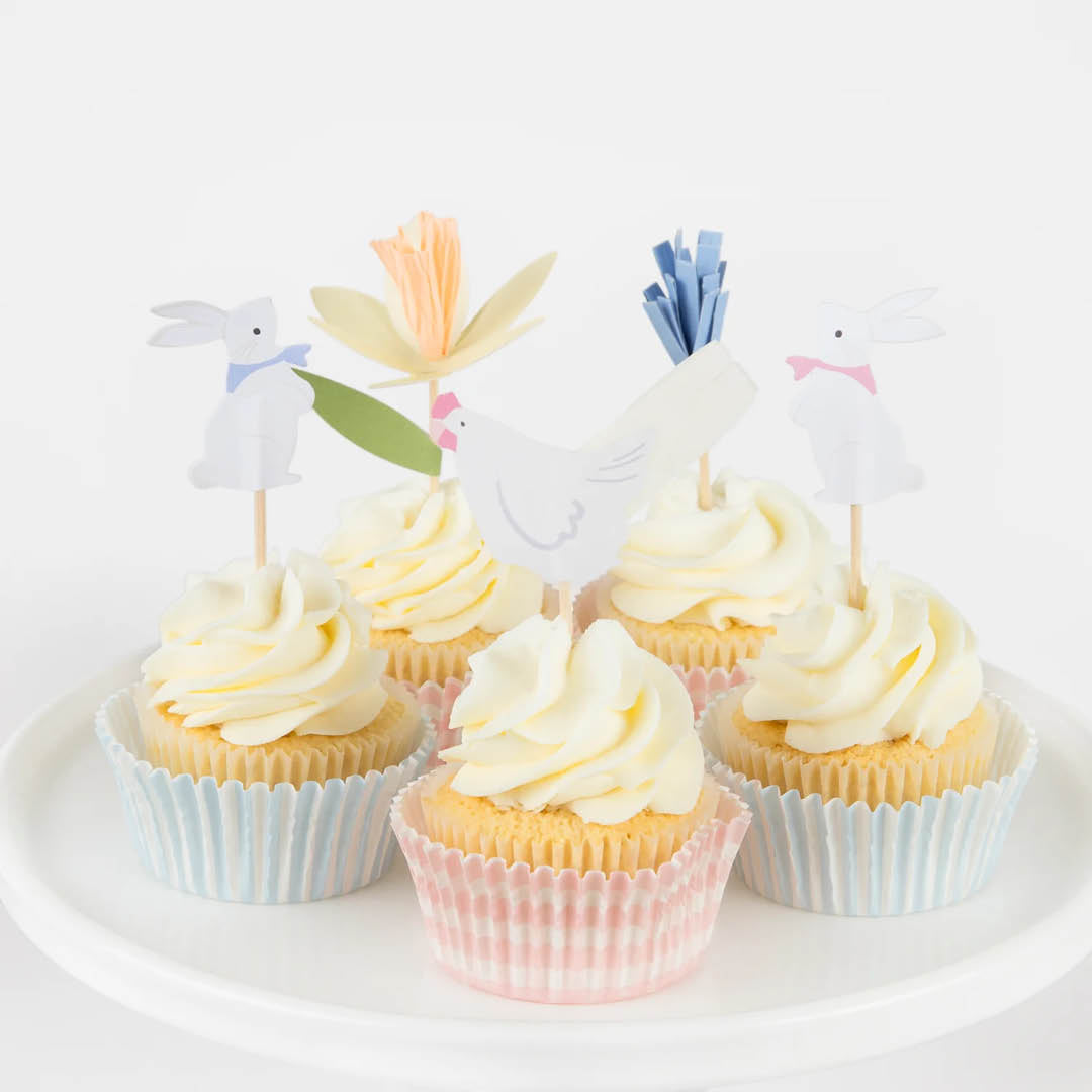 Zes versierde cupcakes met paastoppers van konijntjes, kippen en bloemen op een witte schaal.