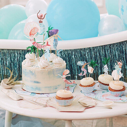 Een feesttafel met een taart en cupcakes versierd met de zeemeerminnen cupcake set en blauwe ballonnen.