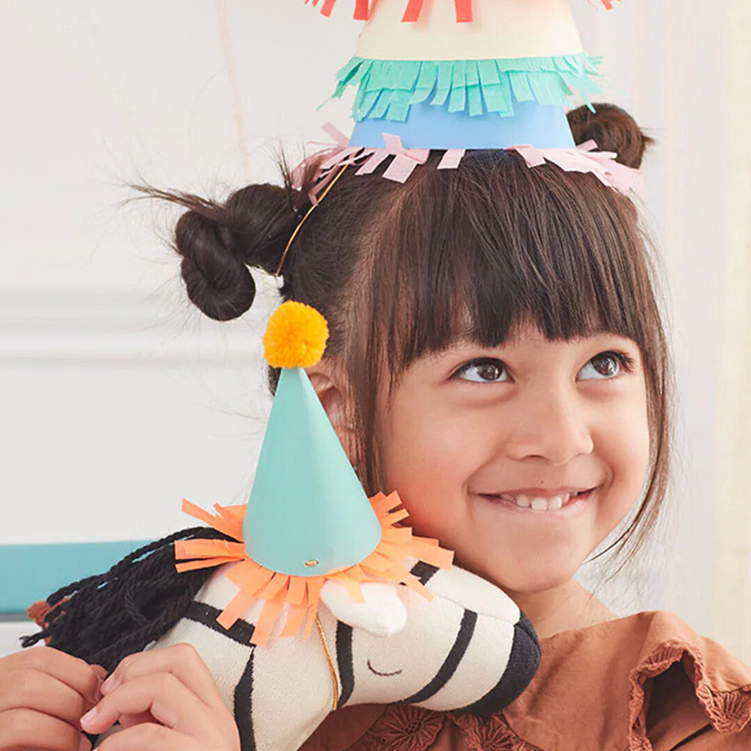 Een lachend kind met een mini feesthoedje op haar hoofd, dat een zebra knuffel met een feesthoedje vasthoudt.