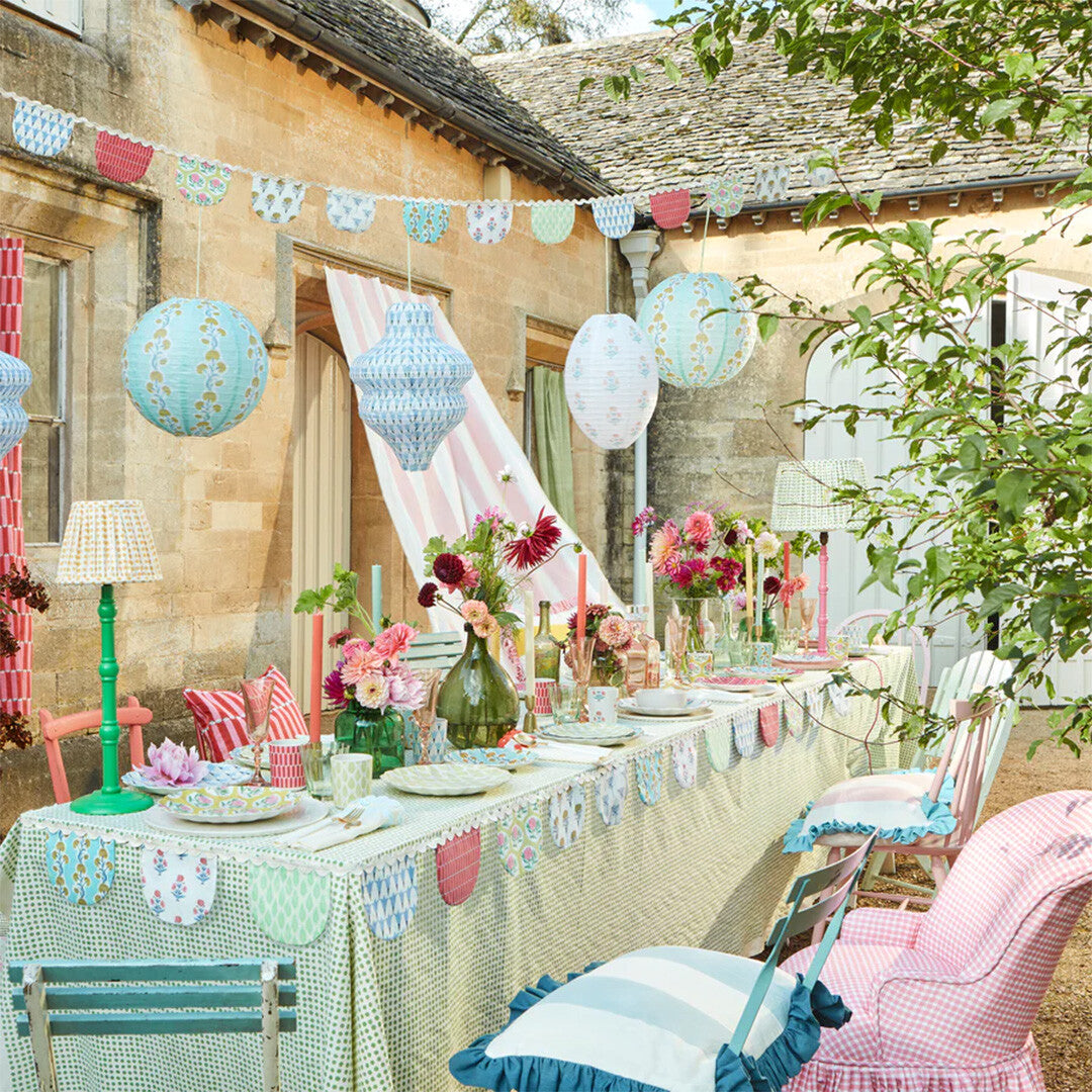 Een feestelijk gedekte tafel buiten met Meri Meri lampionnen, slingers en bloemdecoraties.