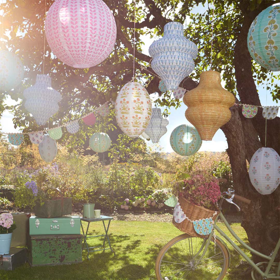Meri Meri papieren lampionnen in diverse kleuren en patronen hangen feestelijk in een zonnige tuin aan bomen.