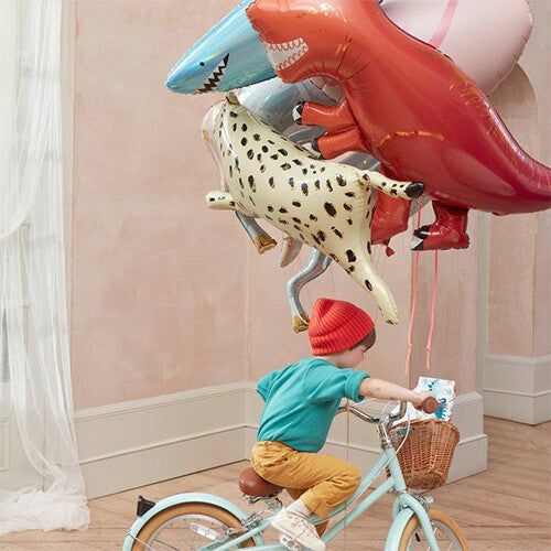 Een kind op een fiets kijkt naar een groep Meri Meri folieballonnen, waaronder een rode T-Rex en een blauwe haai.