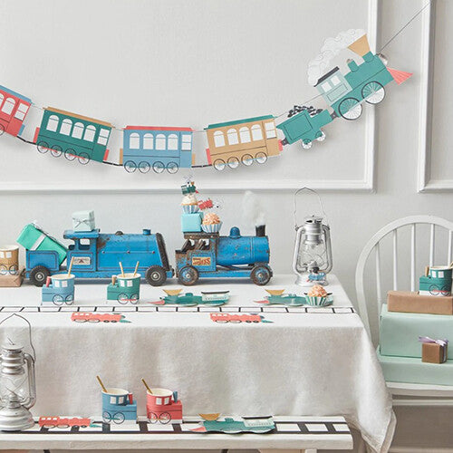 Een feestelijk gedekte tafel met Meri Meri trein thema decoraties, waaronder een slinger en servies.