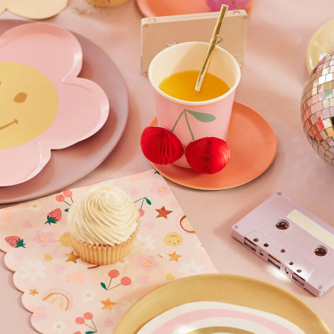 Een Meri Meri papieren servet met vrolijke figuurtjes ligt op een feestelijk gedekte tafel naast een cupcake en een drankje.