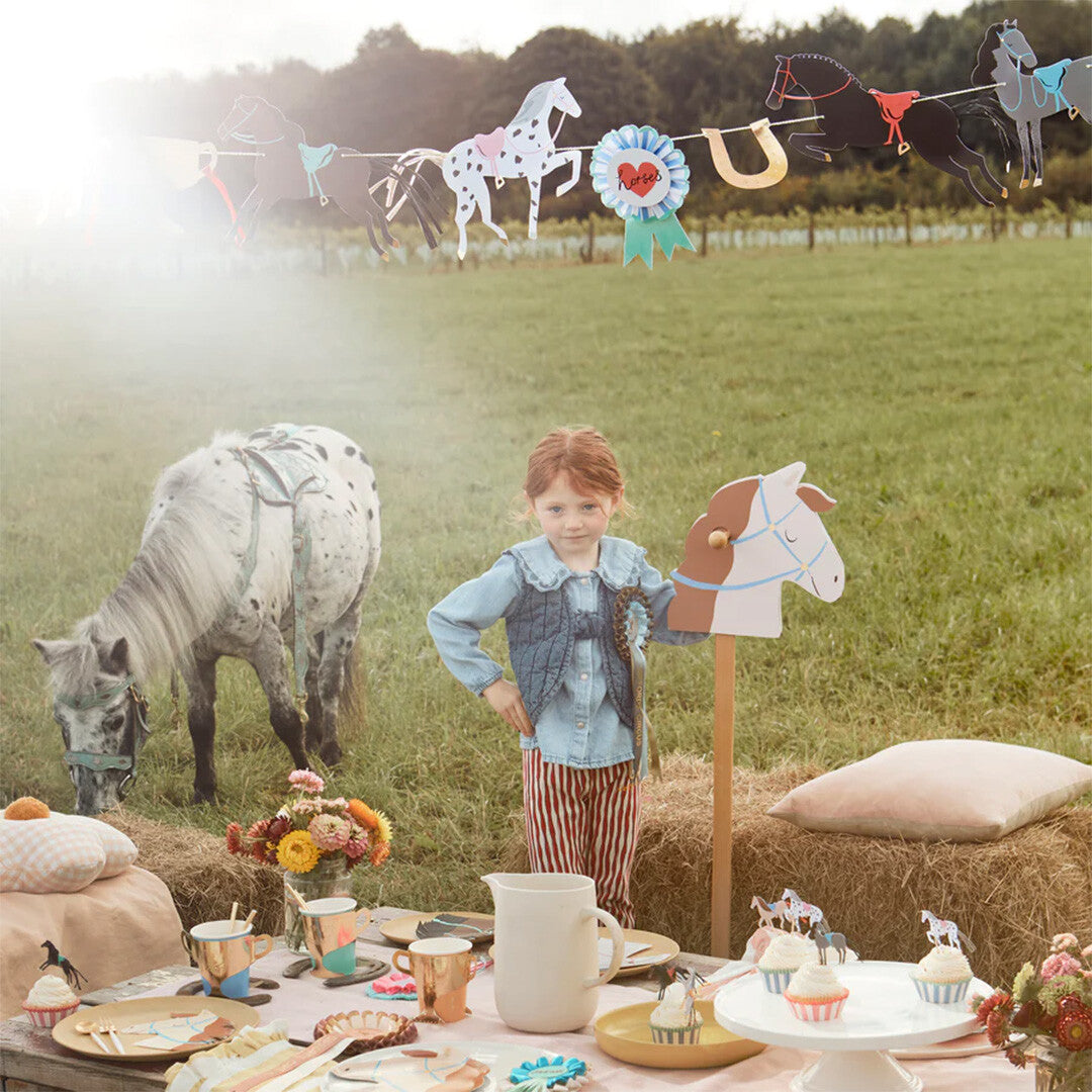 Een meisje bij een pony en een feesttafel, met de Meri Meri paardenslinger als decoratie in een veld.
