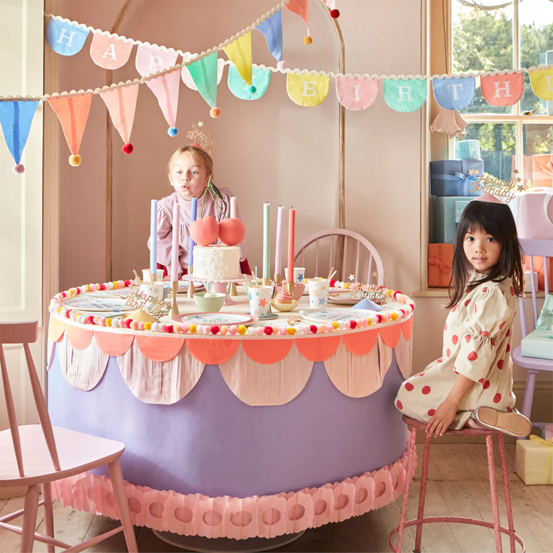 Twee kinderen vieren een verjaardag met een meri meri slinger van vlaggetjes en pompons boven de feesttafel.