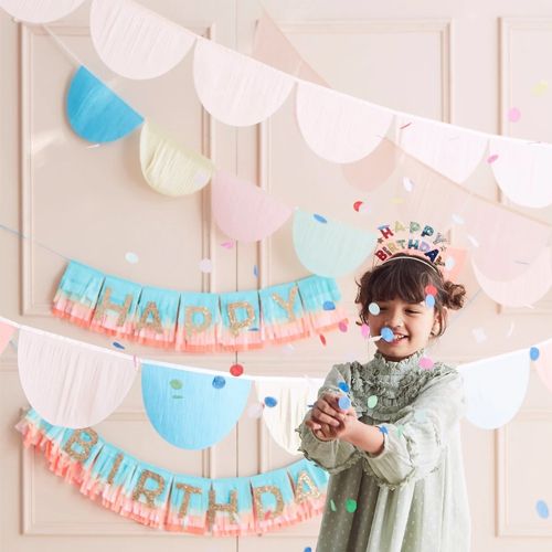 Een lachend meisje gooit confetti voor een achtergrond met Meri Meri regenboog slingers en een Happy Birthday banner.