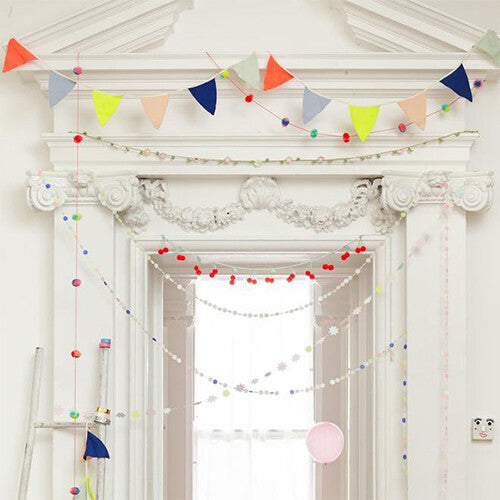 De Meri Meri stoffen slinger met veelkleurige vlaggetjes hangt als feestdecoratie boven een witte deur.
