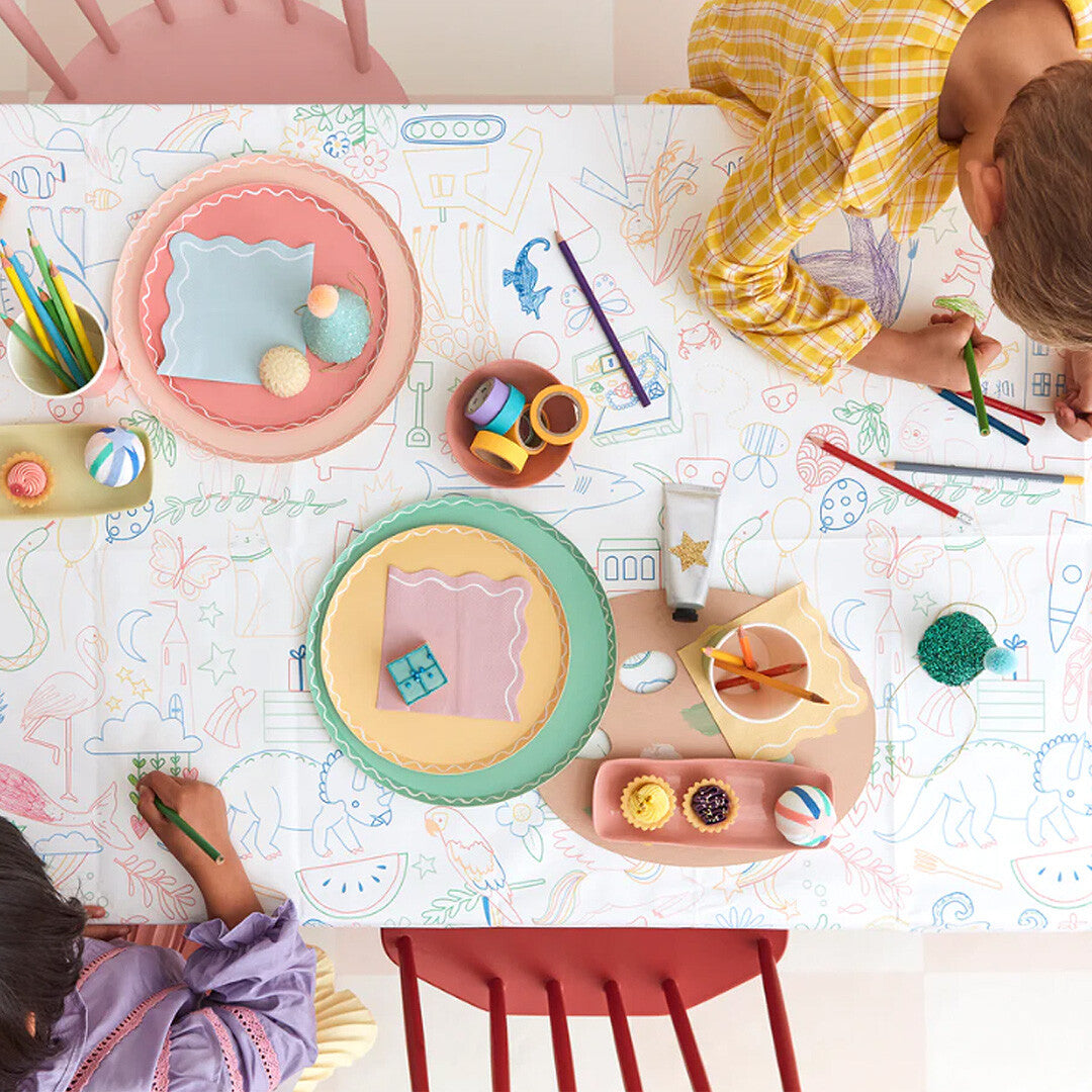 Twee kinderen kleuren op het Meri Meri kleurplaat tafelkleed dat is uitgespreid op een tafel met kleurpotloden en feestdecoraties.