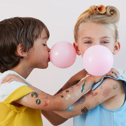 Twee kinderen met Meri Meri tijdelijke tatoeages op hun armen, blazend op roze kauwgomballen.