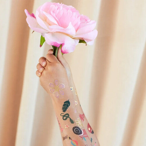 Een kinderarm met diverse tijdelijke tatoeages van bloemen en natuurlijke elementen, die een roze pioenroos vasthoudt.