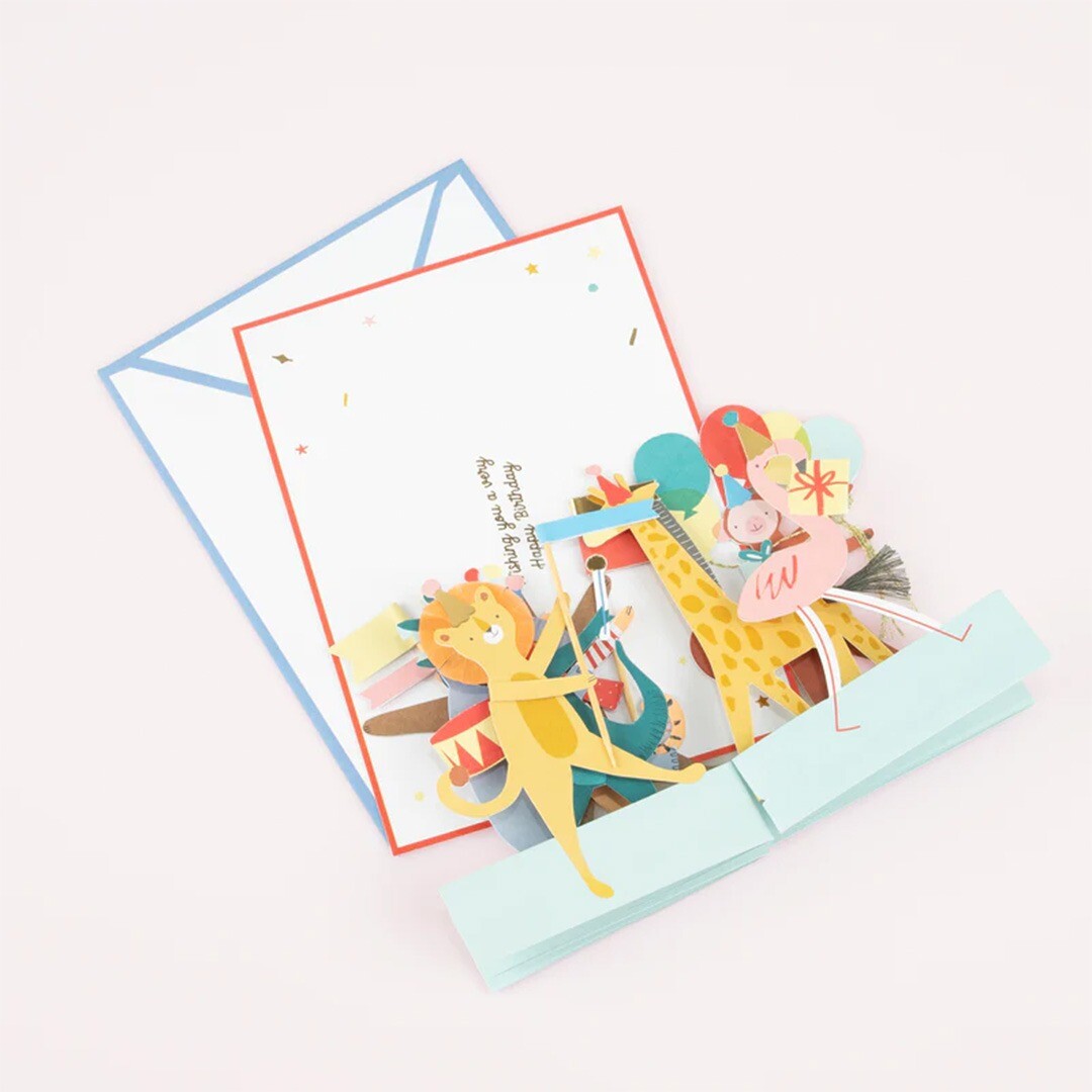 Meri meri verjaardagskaart met pop-up dierenparade en een witte envelop op een lichte ondergrond.