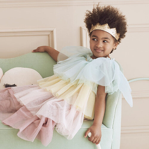 Een kind draagt de Meri Meri regenboogprinses verkleedkleding, zittend op een lichtgroene bank.