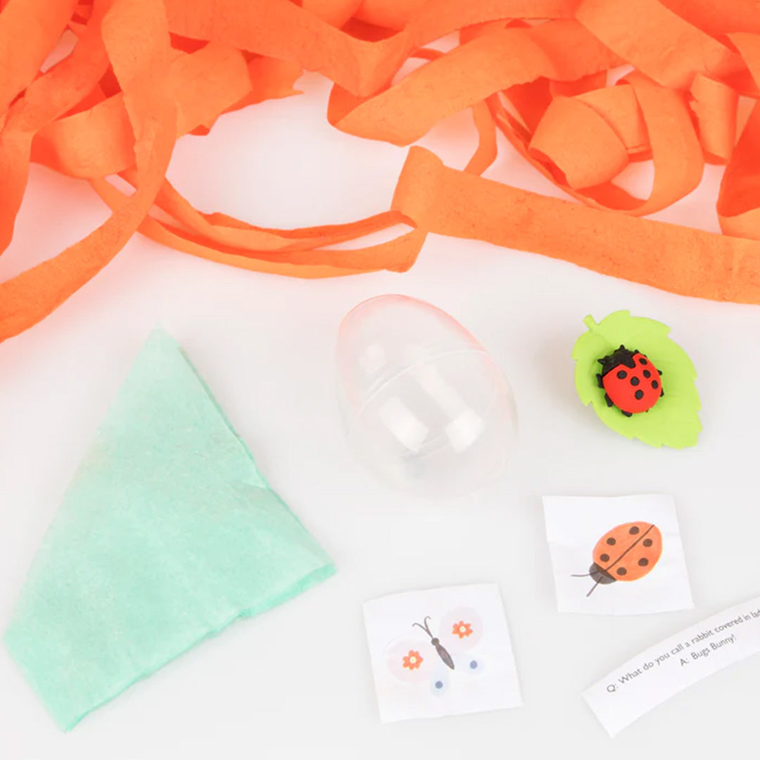 De inhoud van een verrassingsbal uitgestald, met oranje slingers, een plastic ei, een groen doekje, kleine kaartjes en een lieveheersbeestje gum.