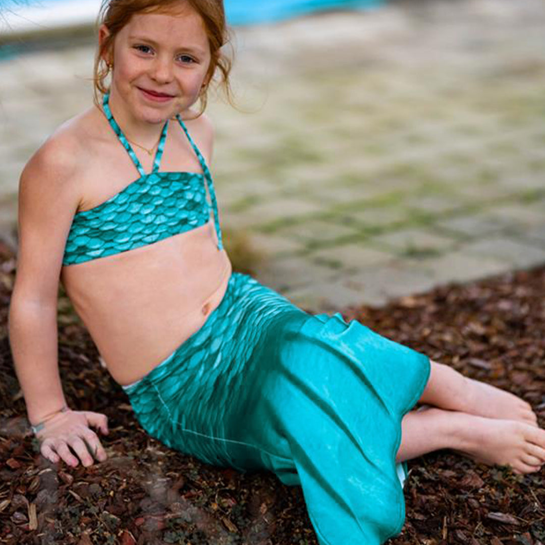 Een meisje zit op de grond en draagt de turquoise zeemeerminstaart en bikinitop.