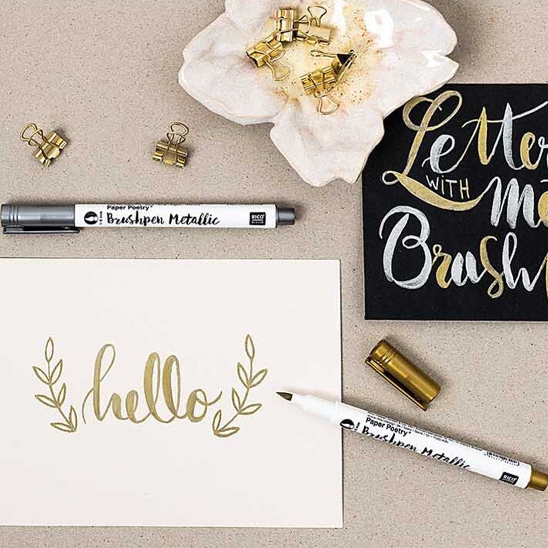 Rico Design metallic brushpens in goud en zilver die 'hello' en 'lettering' schrijven op papier.