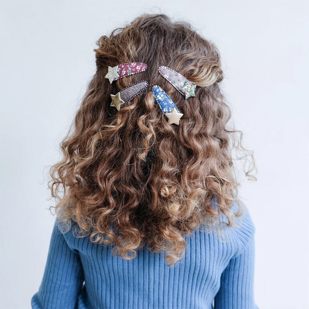 Een kind met krullend haar draagt meerdere Mimi & Lula haarclipjes met bloemen en sterren.