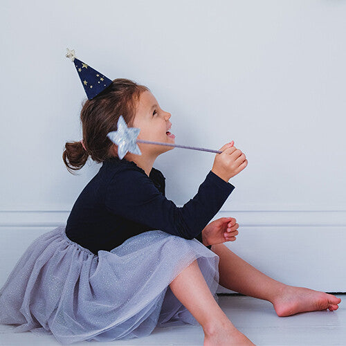 Meisje zit op de grond en speelt met een sterrenstaf, gekleed in de mimi & lula grijze tule kinderrok en een sterrenfeestmuts.