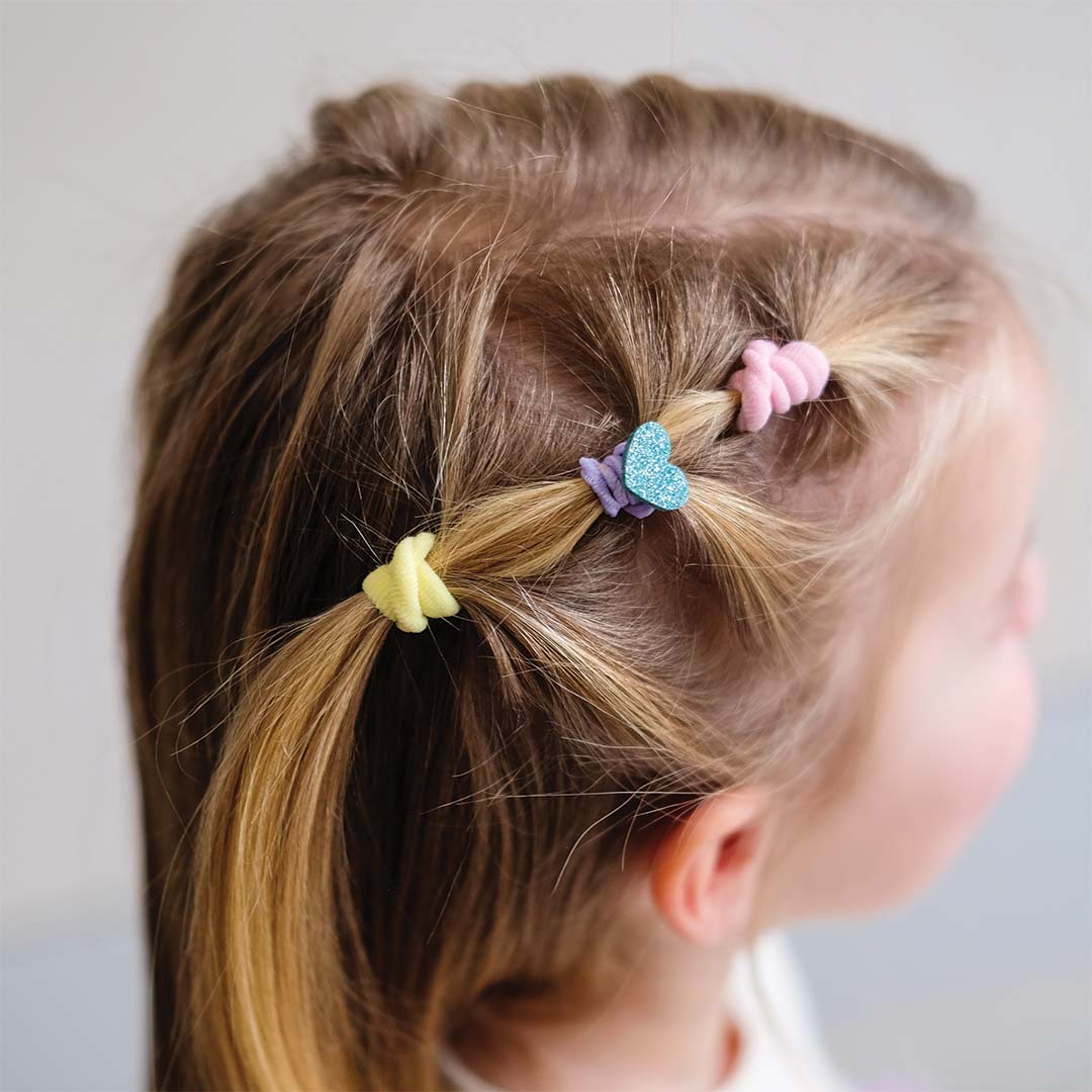 Close-up van een kind met blonde vlechten versierd met kleine pastelkleurige haaraccessoires, waaronder een glitterhartje.