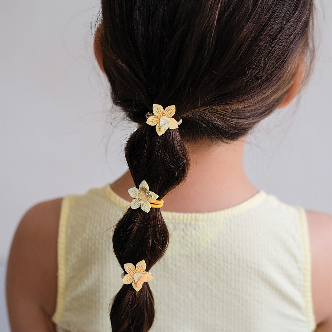 De achterkant van een kind met een vlecht, versierd met mimi & lula haarelastiekjes met gele narcisbloemen.