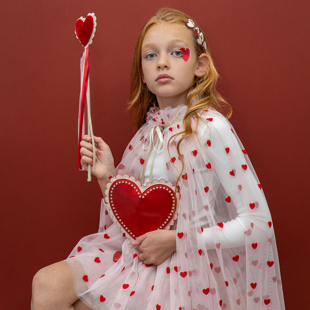 Een kind met rood haar houdt het rode hartvormige schoudertasje en een toverstaf met hart vast.