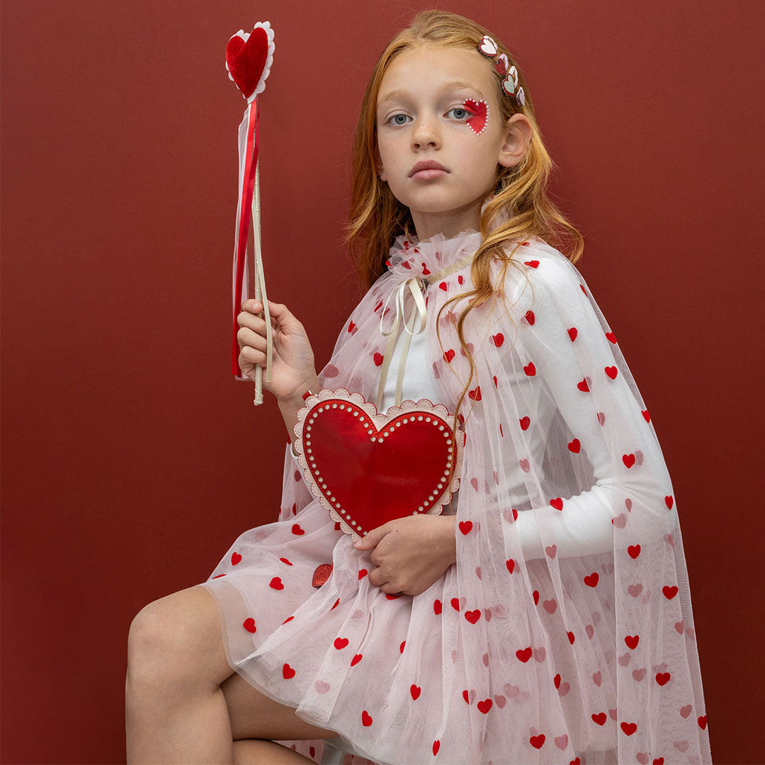 Een meisje met rood haar draagt de roze kinder verkleedcape met harten en houdt een rode hartjestas vast.