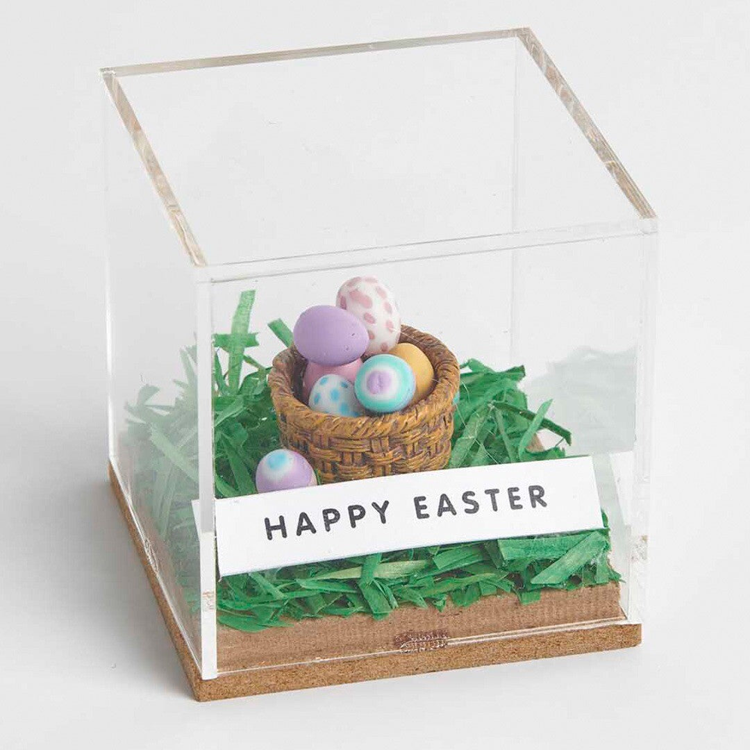 Een miniatuur eiermandje met kleurrijke paaseitjes in een doorzichtige doos met een 'Happy Easter' bordje.