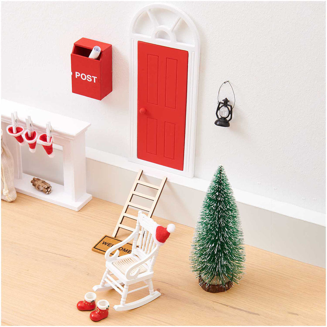 Miniatuur houten schommelstoel met kerstmuts in een kerst-poppenhuisdecor met rode deur, kerstboom en laarsjes.