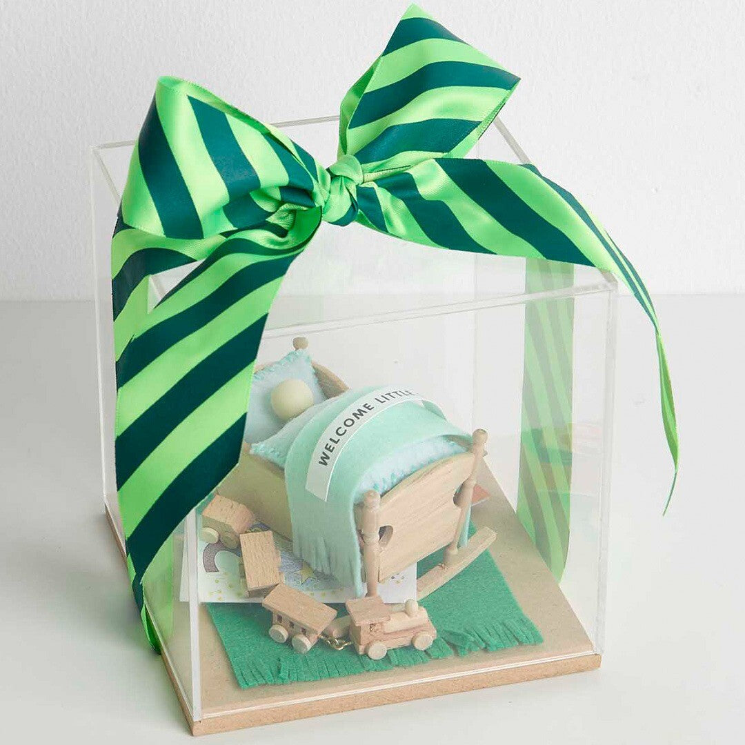 Een Rico Design miniatuur houten wiegje en andere poppenhuisaccessoires in een transparante geschenkdoos met een groene strik.