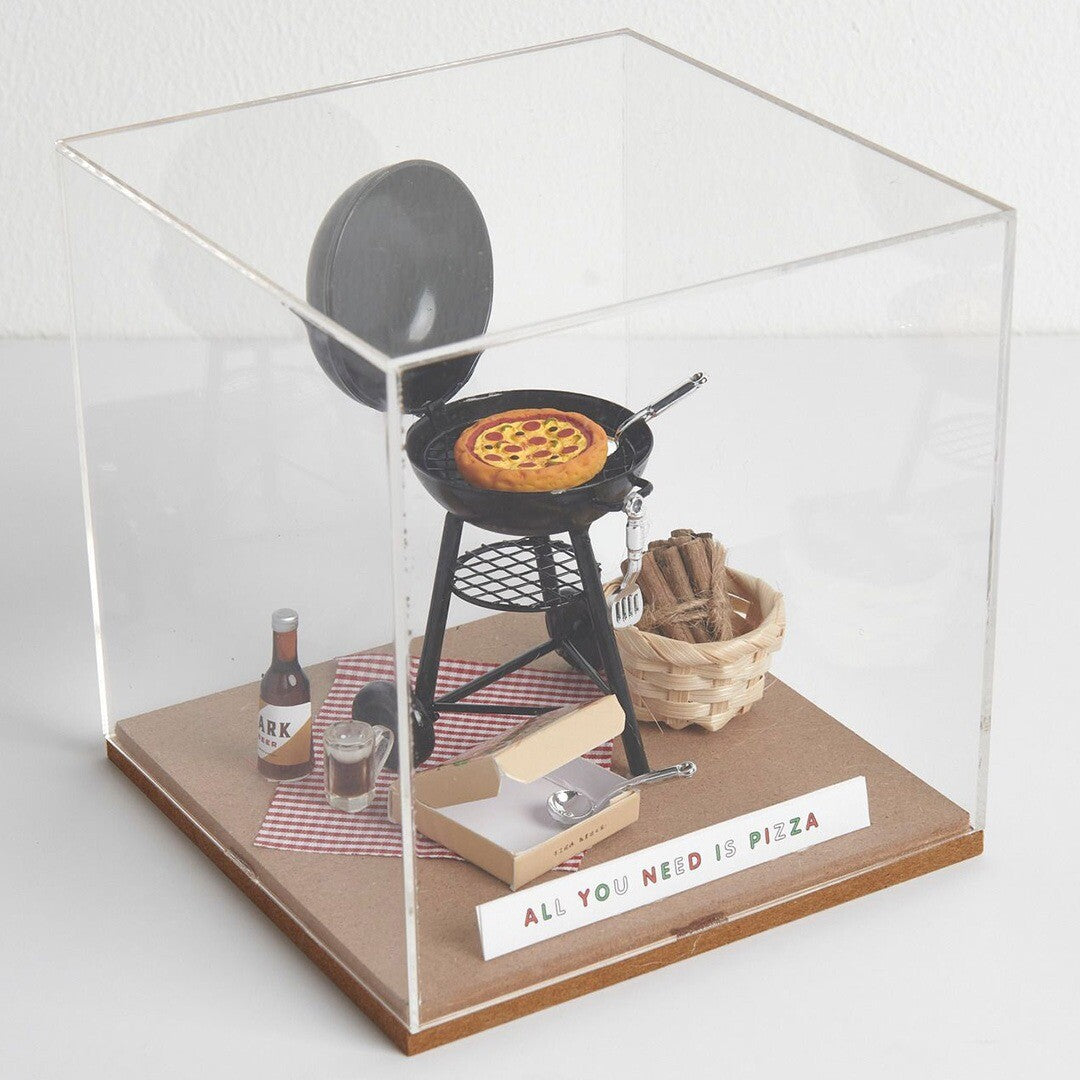 Miniatuur diorama van een barbecue met pizza, een mandje en een picknickdeken in een vitrine.