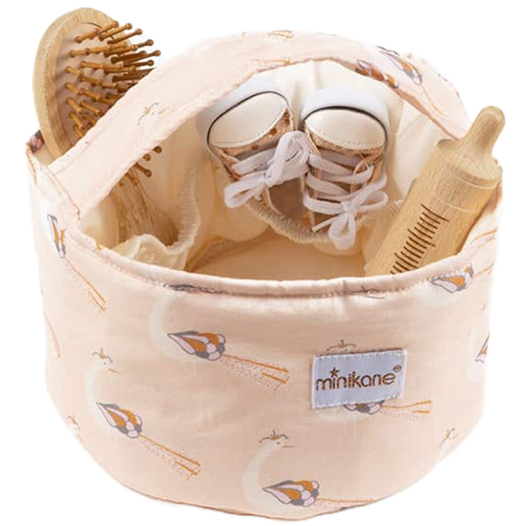 minikane beige stoffen opbergmand met pauwenpatroon gevuld met een houten borstel, poppenschoentjes en een houten fles.