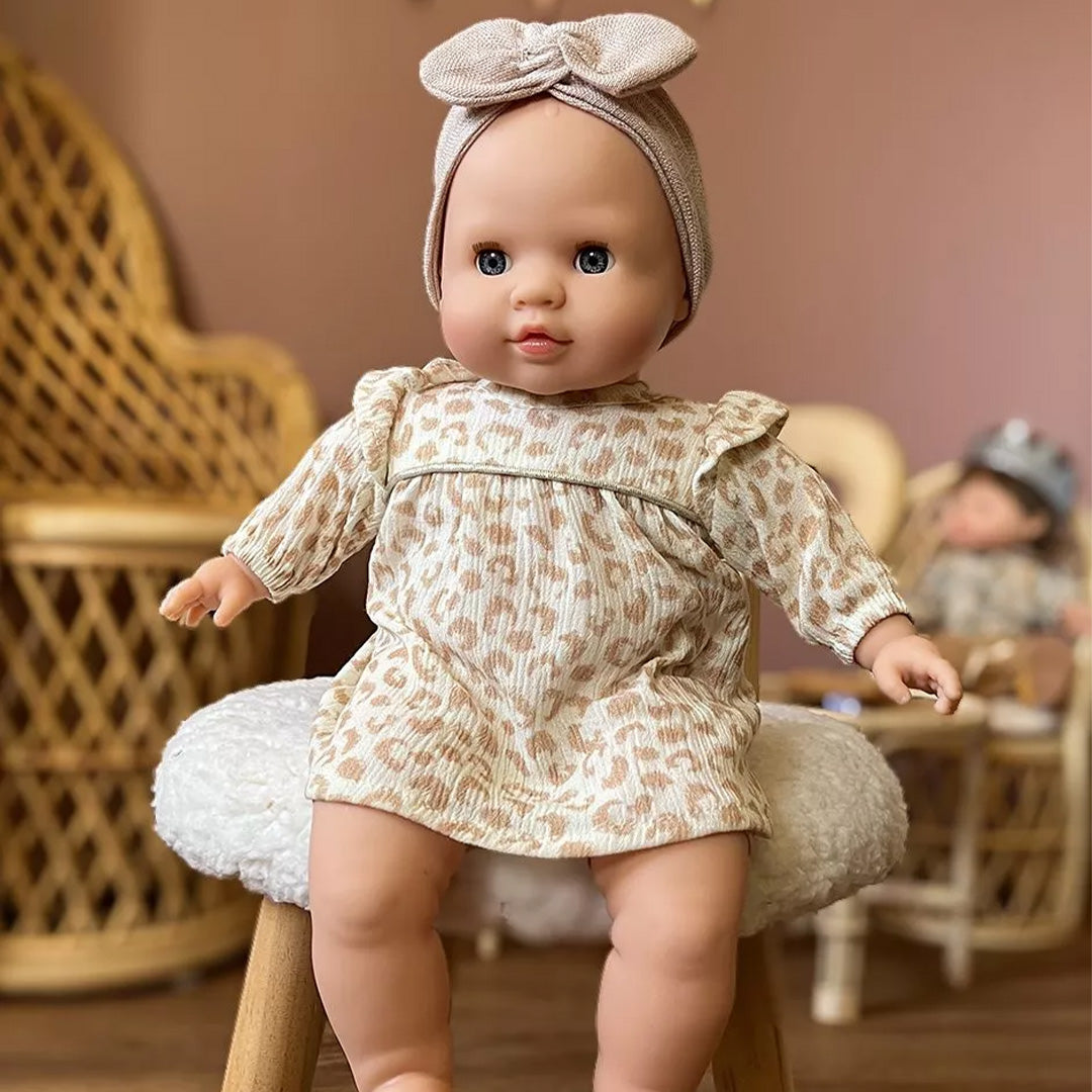 minikane babypop zit op een krukje, gekleed in een beige jurkje met print en een hoofdband.