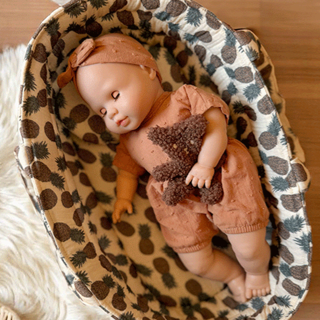 minikane babypop slaapt in een mandje, gekleed in een bruin pakje en met een teddybeer.